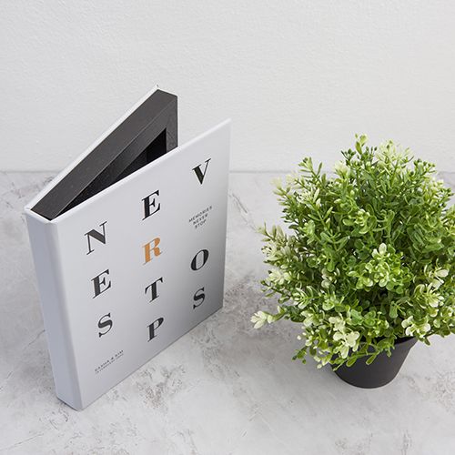 Weißes Buch mit schwarzem Typografietitel und kleiner grüner Topfpflanze auf grauer Oberfläche.