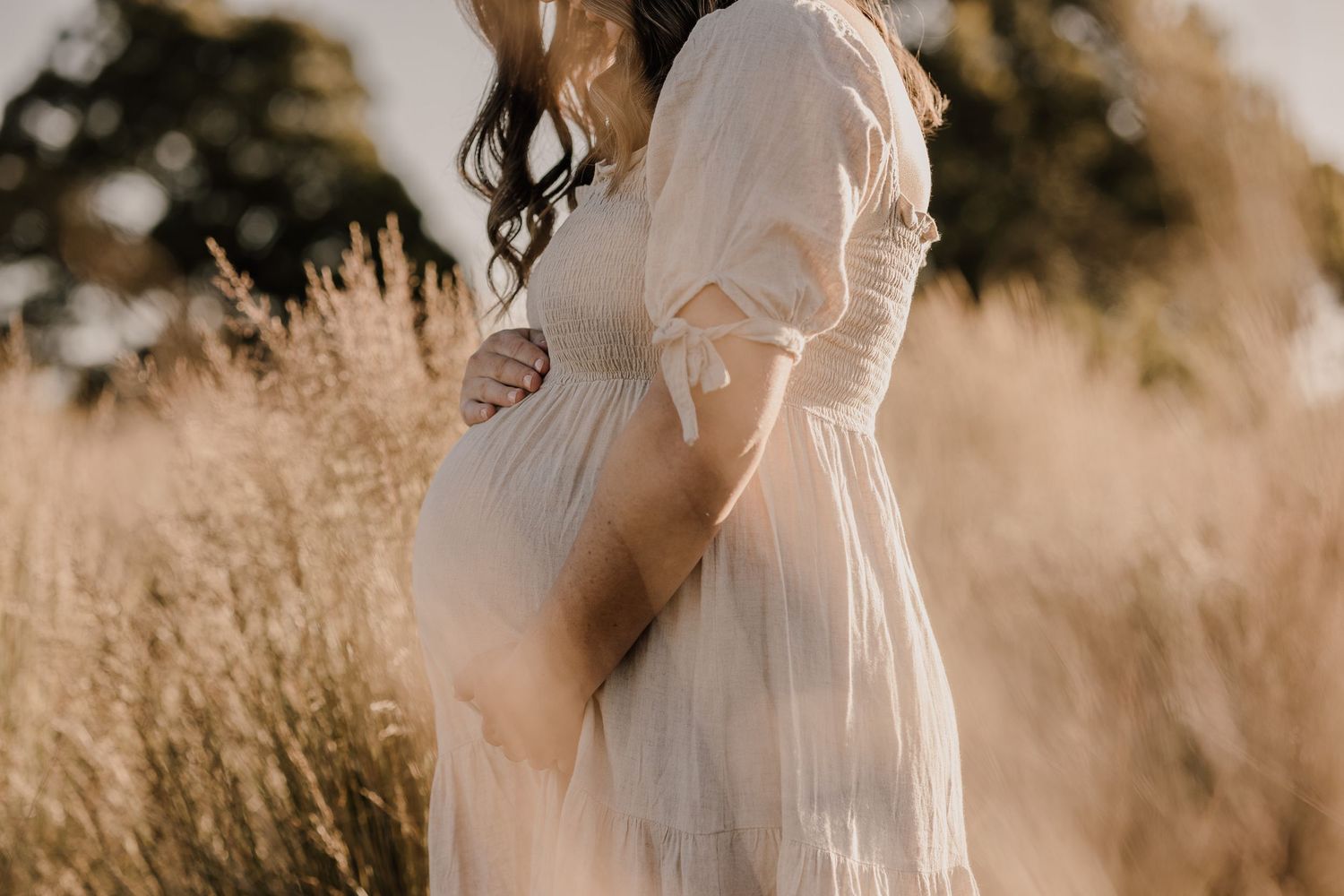 Side view of an expectant mother in a flowing white dress standing in golden sunlit field.