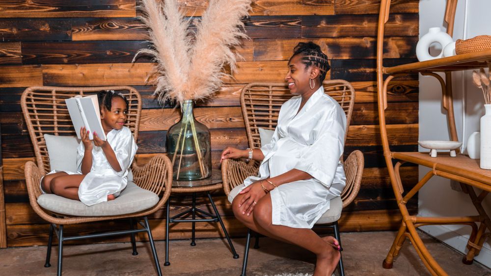 Two people in white outfits sit in modern chairs in a rustic wooden cabin setting with pampas grass decor.