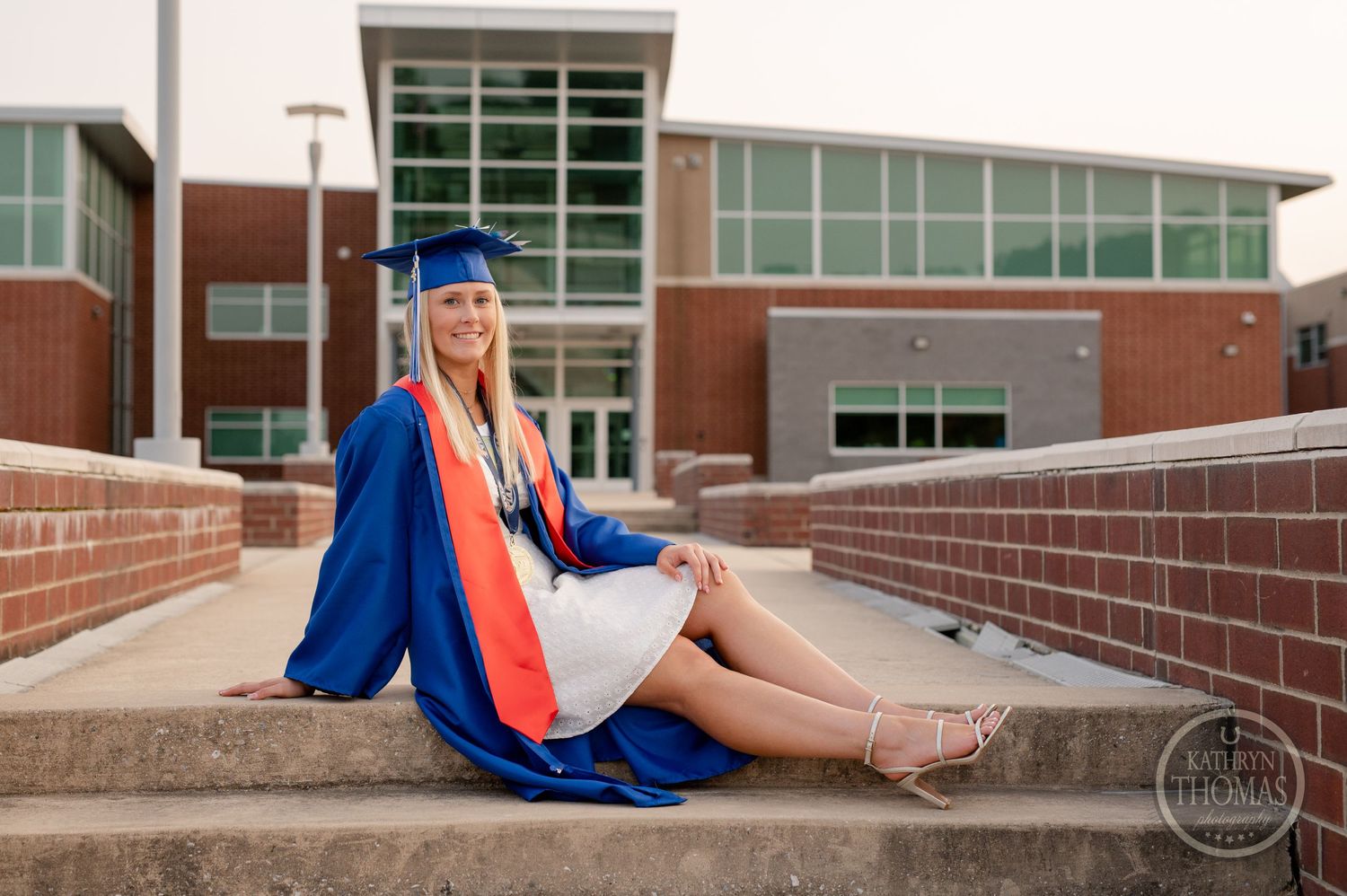 Graduate in traditional blue gown sits on concrete steps outside modern school building.