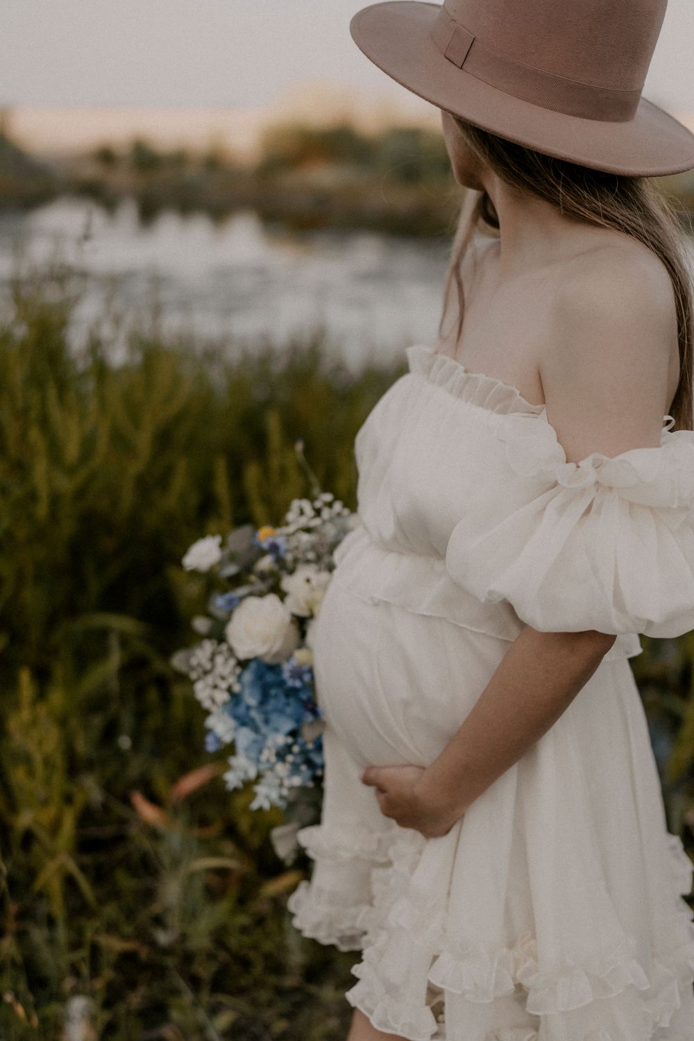 A person in a white dress and brown hat holds a blue and white bouquet against a coastal backdrop.