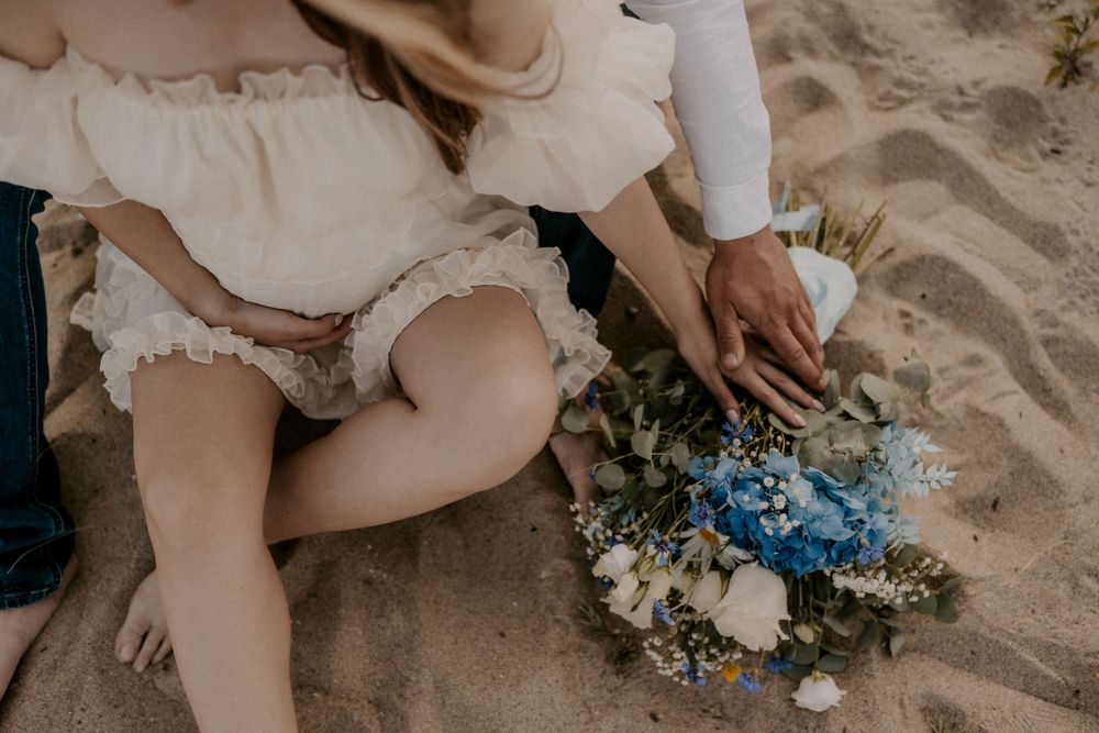 A delicate blue and white bouquet rests on sandy beach near flowing white fabric.