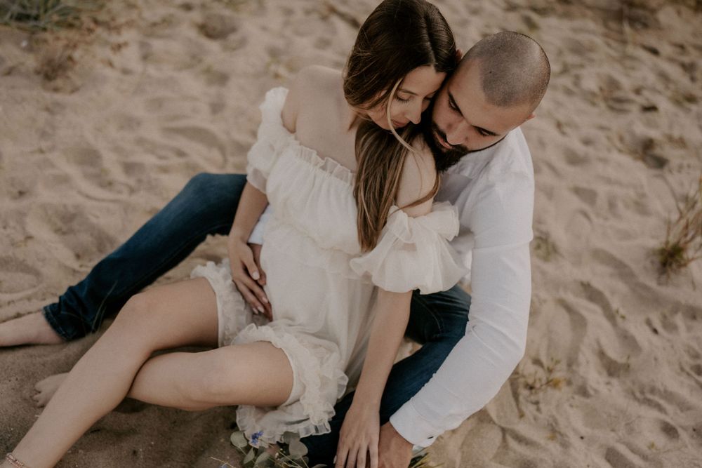 A romantic beach photoshoot featuring a couple sitting together on sandy shores at sunset.