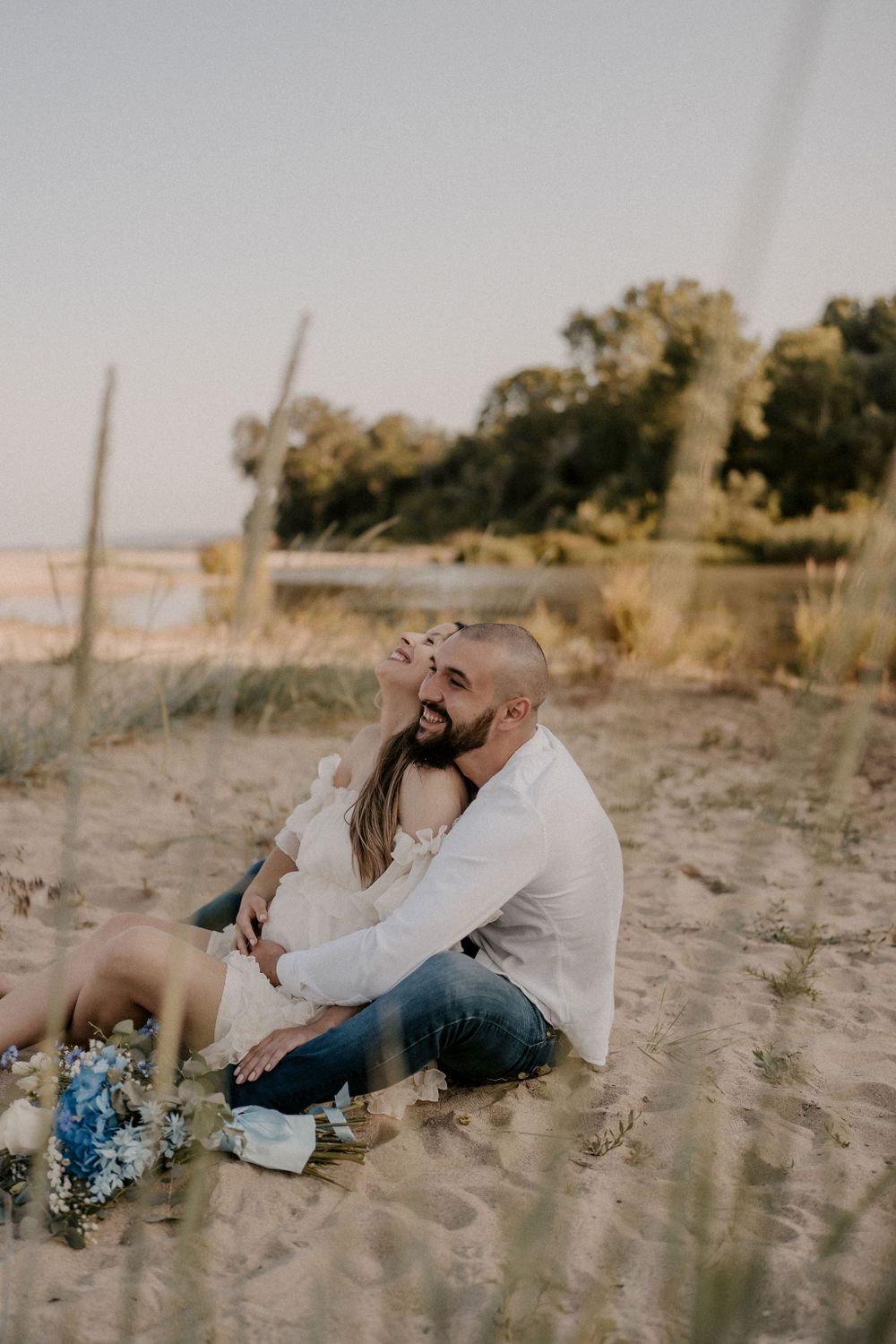 Couple relaxing on sandy terrain with blue flowers beside them during a sunset photoshoot.