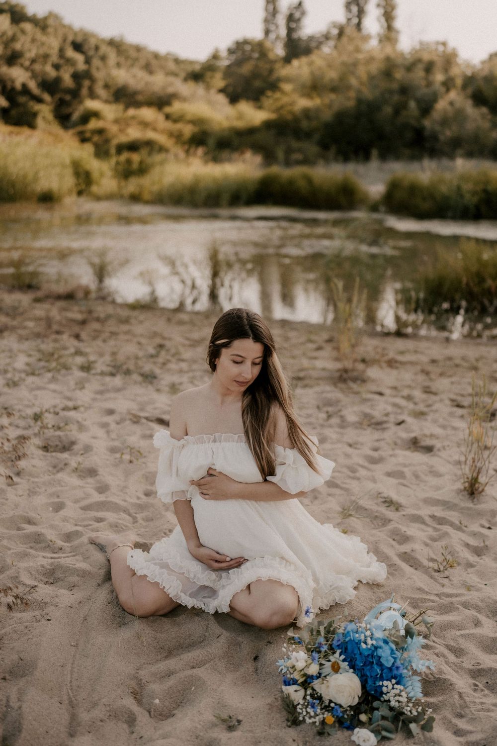 Woman in white dress poses with blue and white floral bouquet on sandy beach.