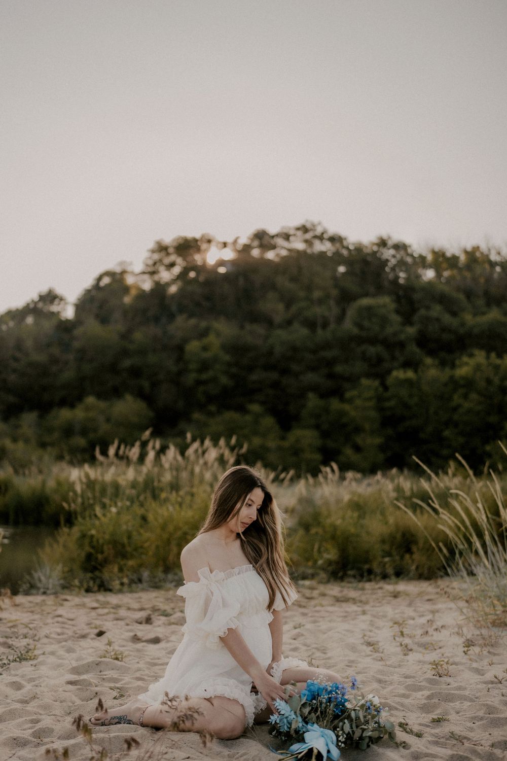 Woman in white dress sits on sandy beach with blue flower bouquet at dusk.