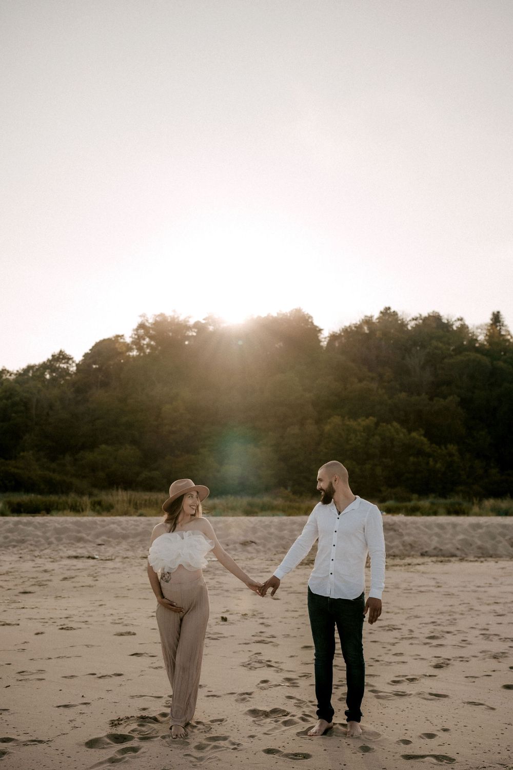 A couple holds hands while walking on sandy beach during golden hour with trees in background.