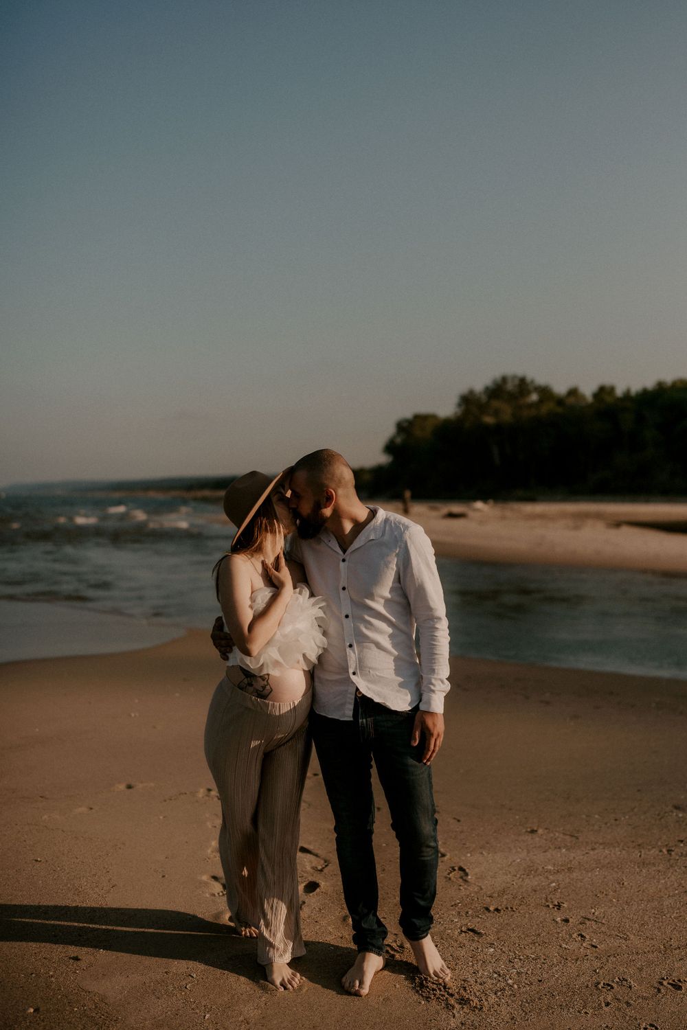A couple shares a romantic moment on the beach with ocean waves and trees in background.