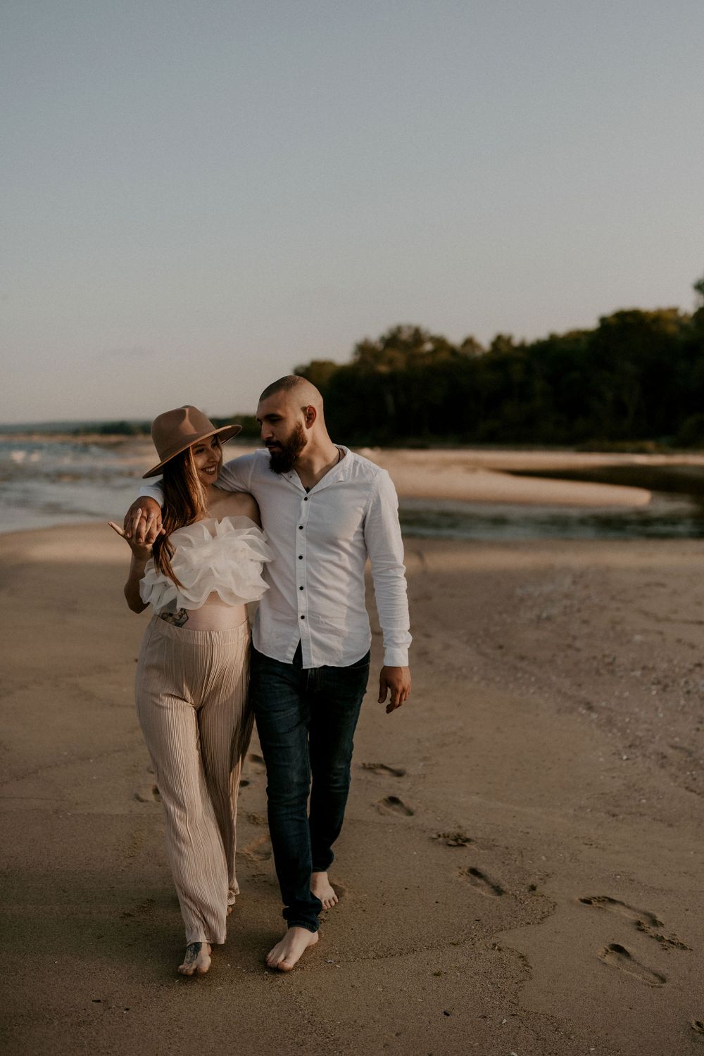 A couple walks barefoot along the shoreline during a warm sunset.