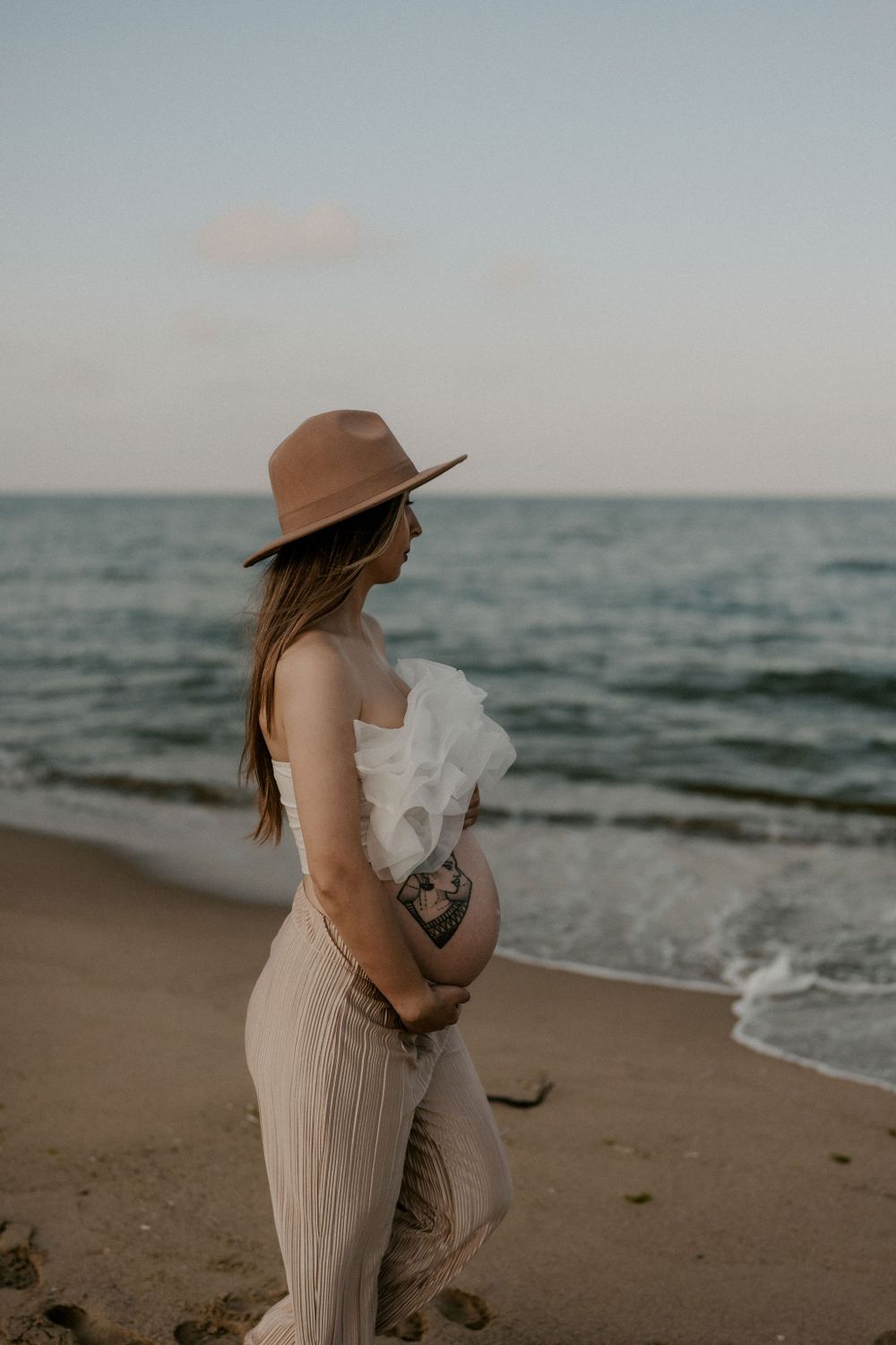 Person in a wide-brimmed hat and flowing dress holds flowers on a peaceful beach.