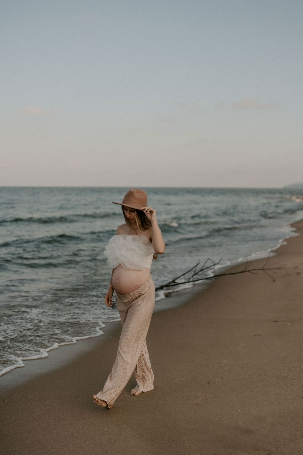 Figure in beige clothing and sun hat strolls along the shoreline at dusk.