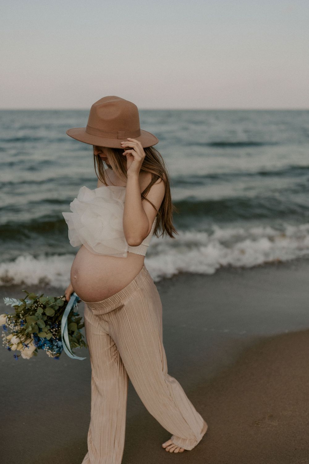 Person in flowing dress and hat carries blue and white flowers on the beach.