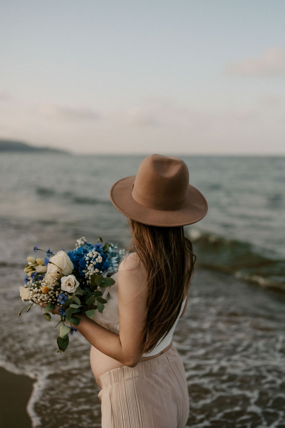 Beachside portrait with flowing dress, hat, and blue floral bouquet.