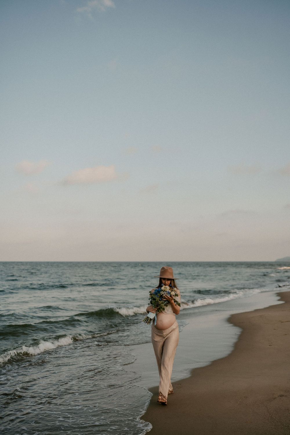 Distant figure walks along empty beach carrying flowers at sunset.