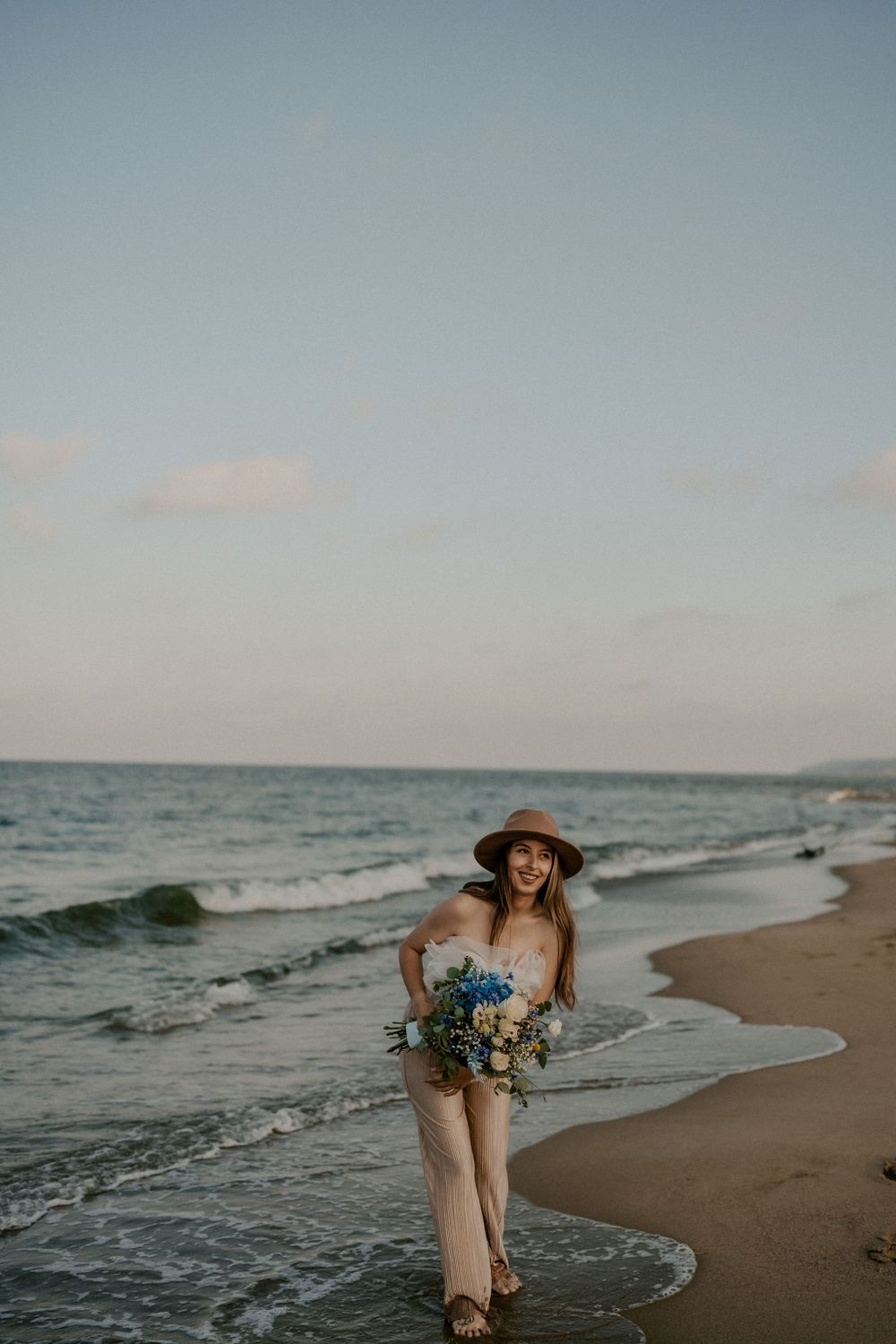 Romantic maternity photoshoot on a beach at sunset with a bouquet of blue and white flowers.