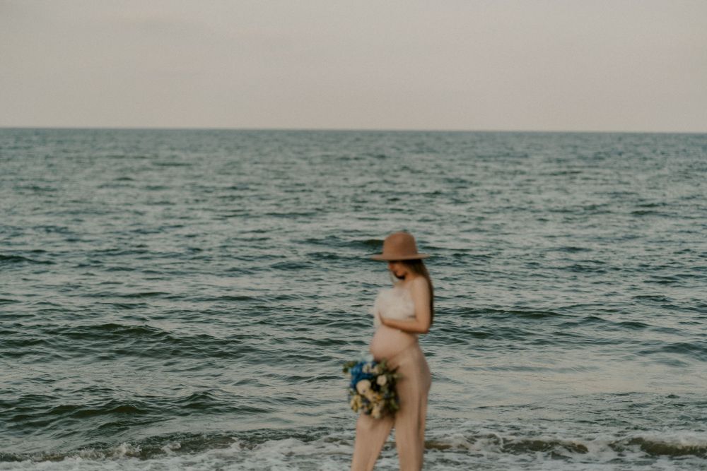 Expecting mother in a flowing dress walks along the shoreline at dusk holding a floral bouquet.