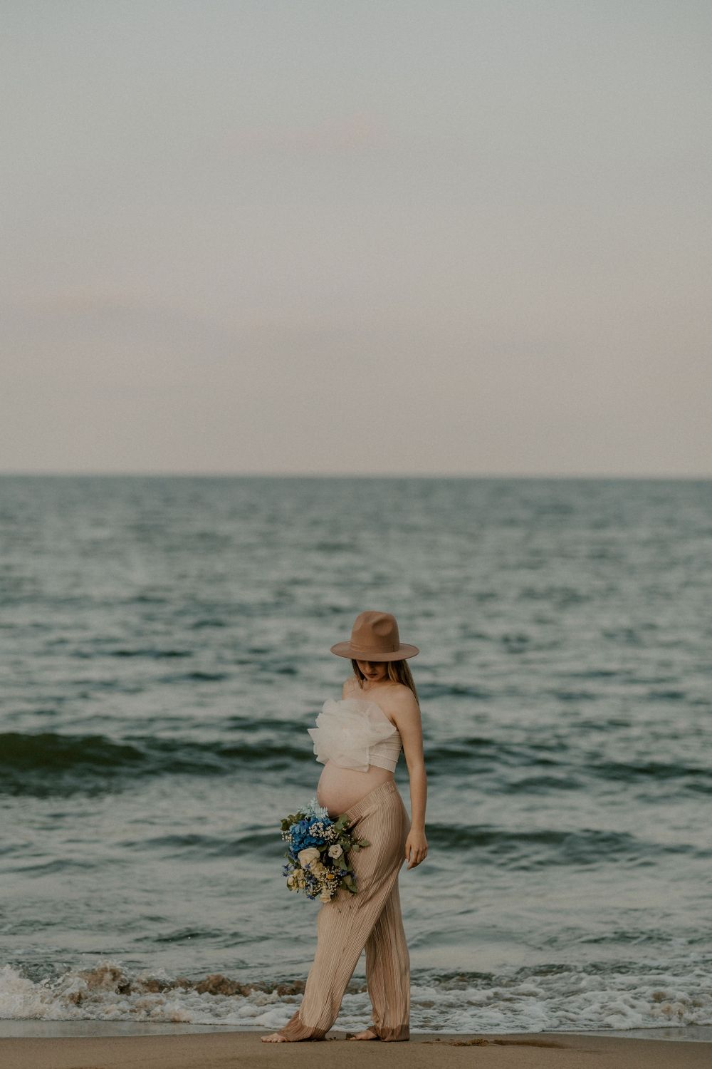 Pregnant woman in beige dress poses gracefully by the ocean waves during sunset.