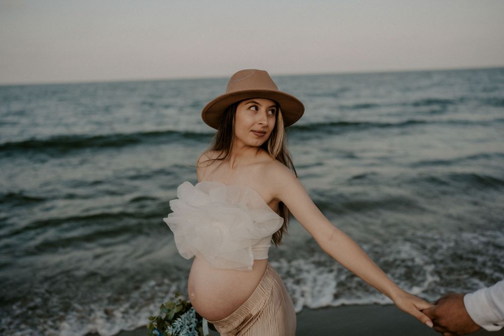 Expectant mother in sun hat and flowing dress poses with flowers by the ocean.