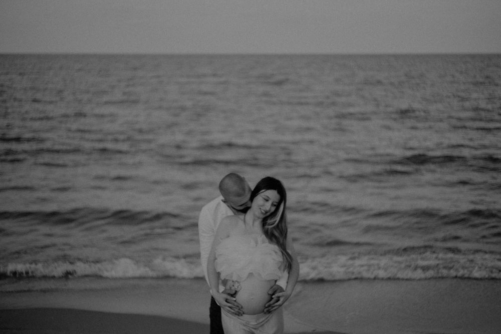 Black and white maternity photo of couple embracing by the ocean.