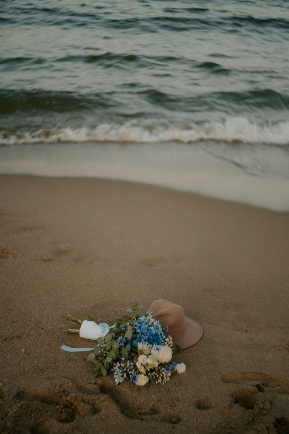 Blue and white floral arrangement lies on sandy beach with ocean waves in background.