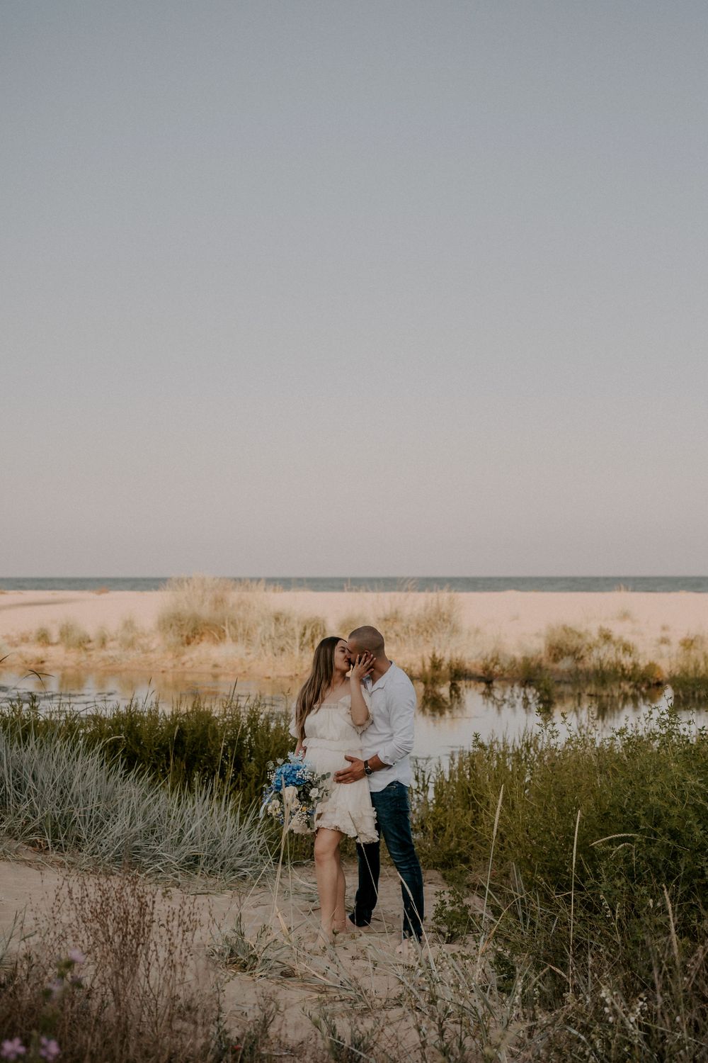 A romantic couple embraces among coastal dunes and beach grass at sunset.