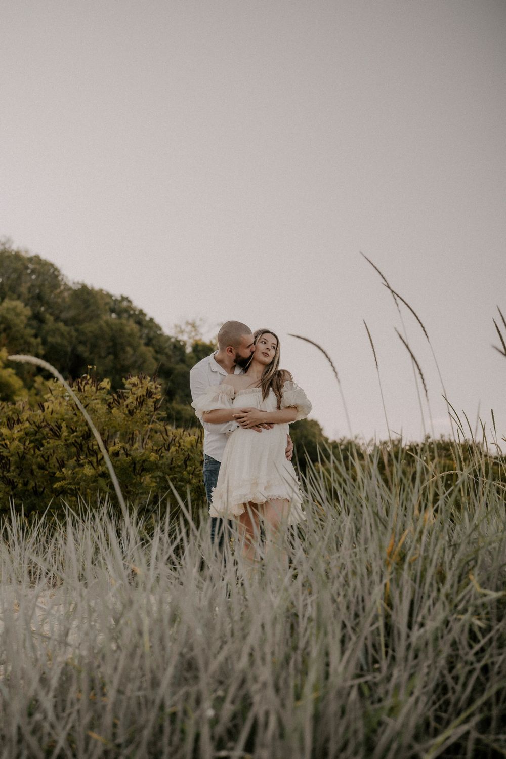 A loving couple shares a tender moment among tall beach grass at dusk.