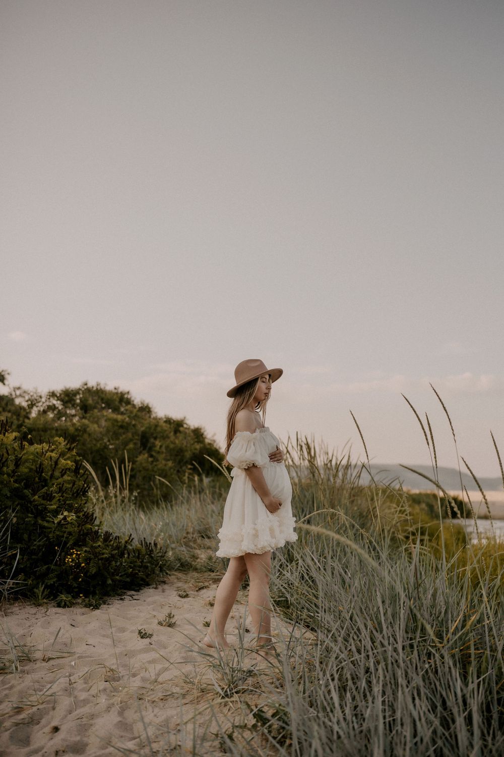 A figure in a flowing white dress stands among coastal dunes at sunset.