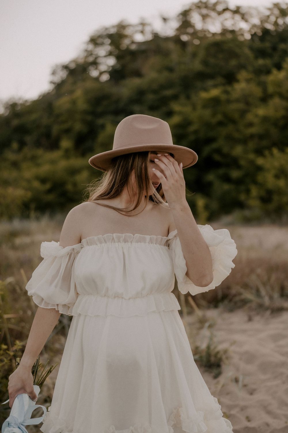A flowing white off-shoulder dress with ruffles is worn on a beach with natural foliage in the background.