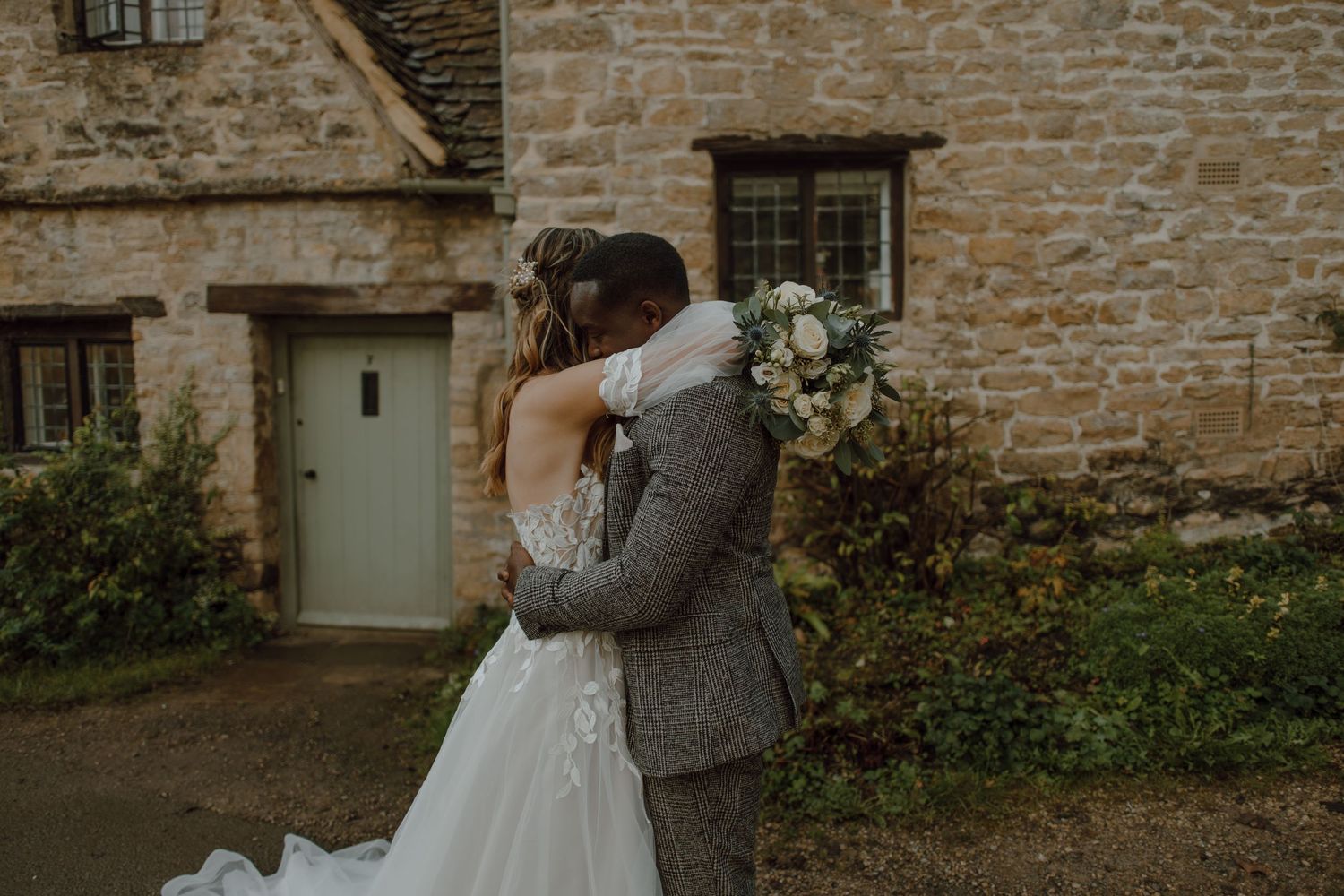 A tender wedding moment captured against a rustic stone wall with white floral arrangements.