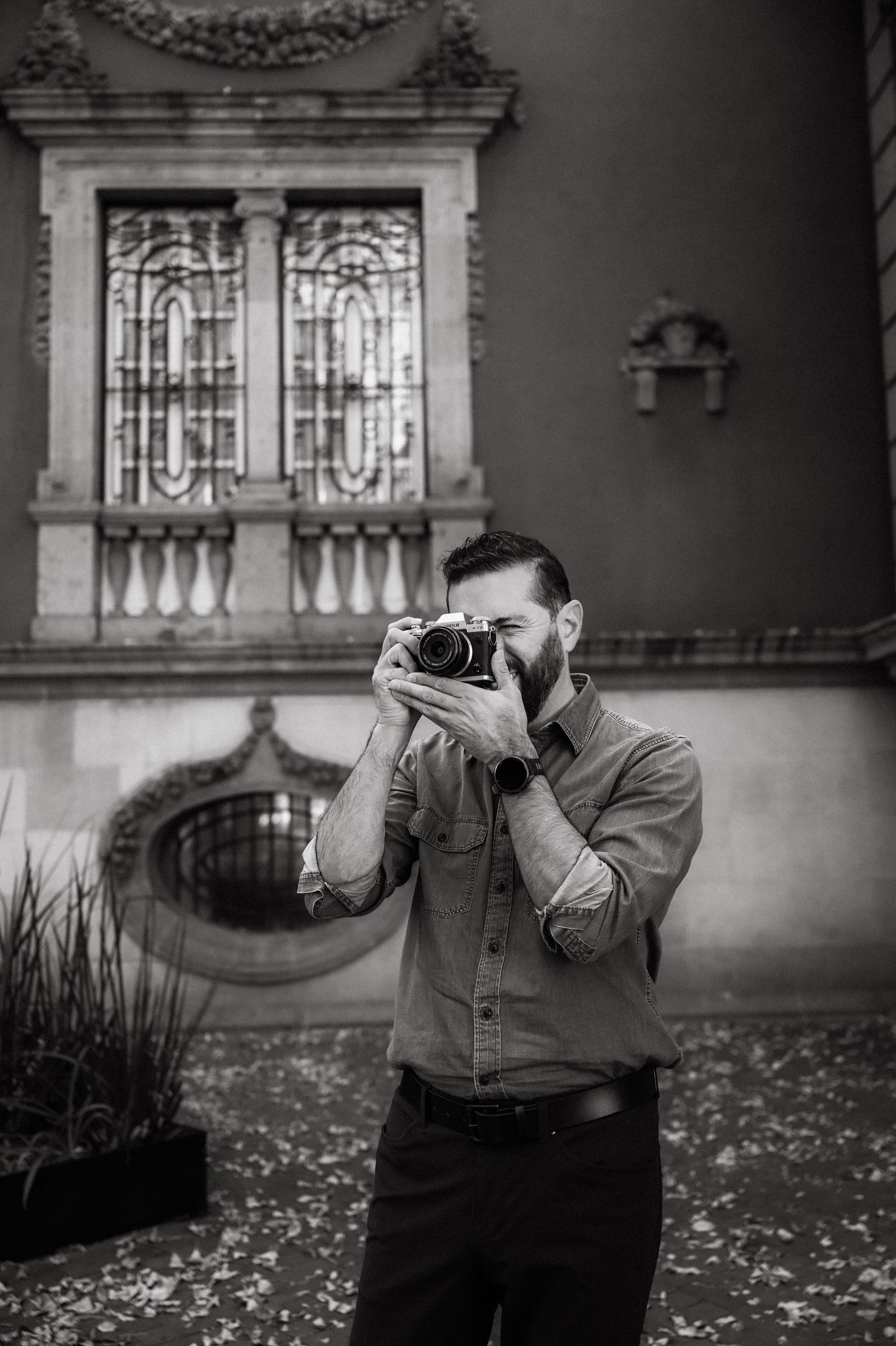 A photographer in casual attire takes pictures in front of a historic building with fallen autumn leaves.