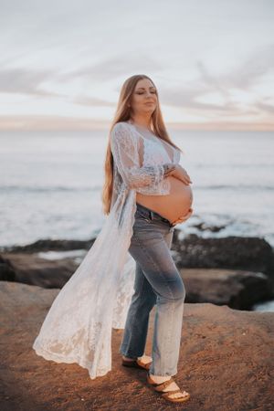 Pregnant woman in white lace kimono and jeans standing on rocky beach during golden hour.