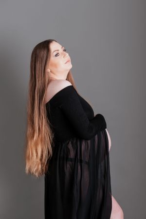 Maternity photo of a woman in a sheer black dress against a grey studio background.