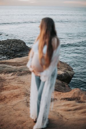 Silhouetted figure in white lace robe standing on ocean cliff during sunset.