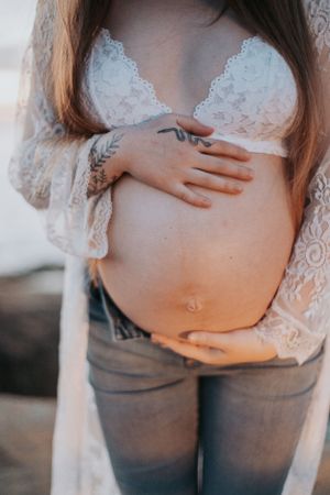 Close-up of pregnant belly with hands in white lace and denim against ocean backdrop.