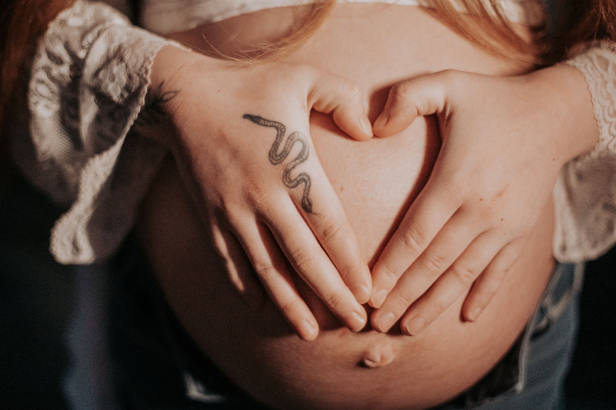 Close-up of hands forming a heart shape over a pregnant belly in soft lighting.