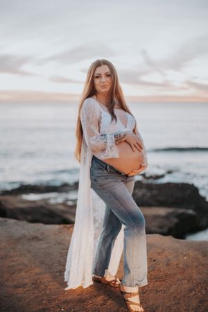 Maternity photo of an expectant mother in a lace robe and jeans posing on coastal rocks at sunset.