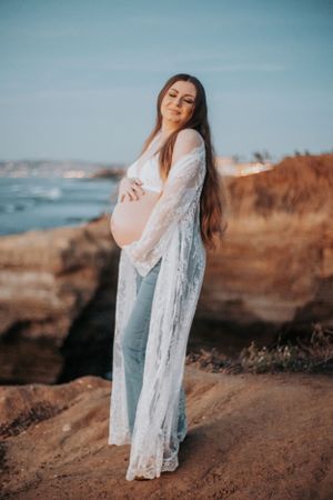 Maternity portrait on coastal cliffs with woman in flowing white lace robe against blue sky.