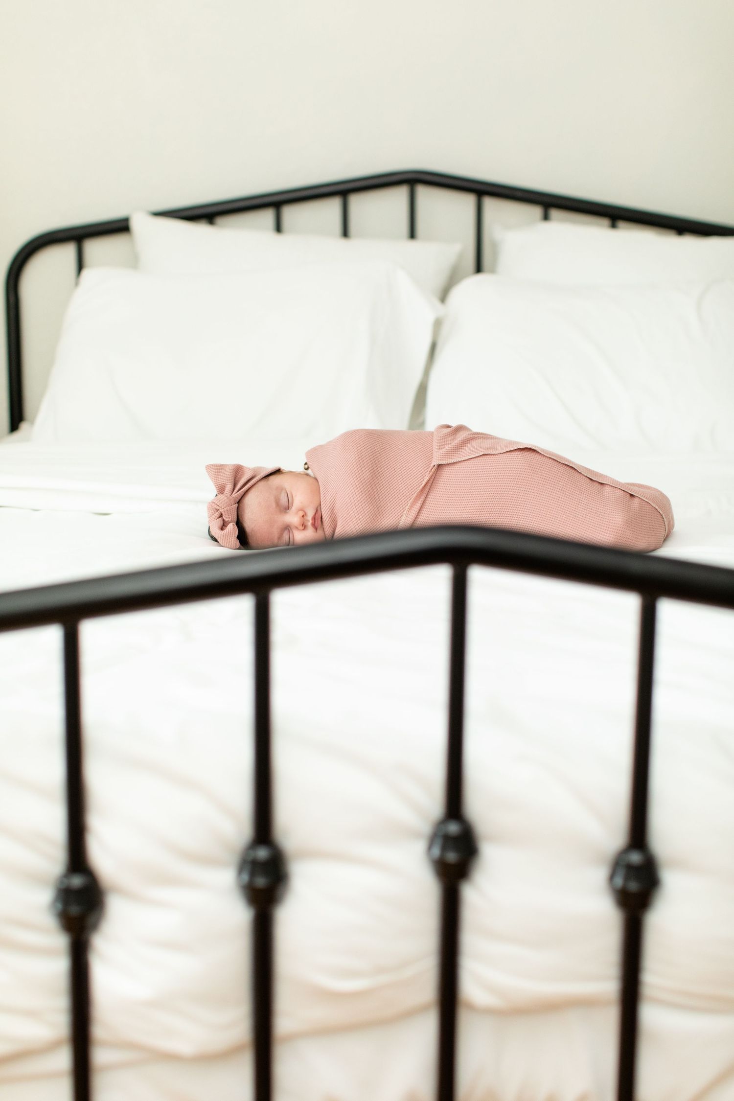 Sleeping baby wrapped in pink swaddle rests on white bed with black metal frame.