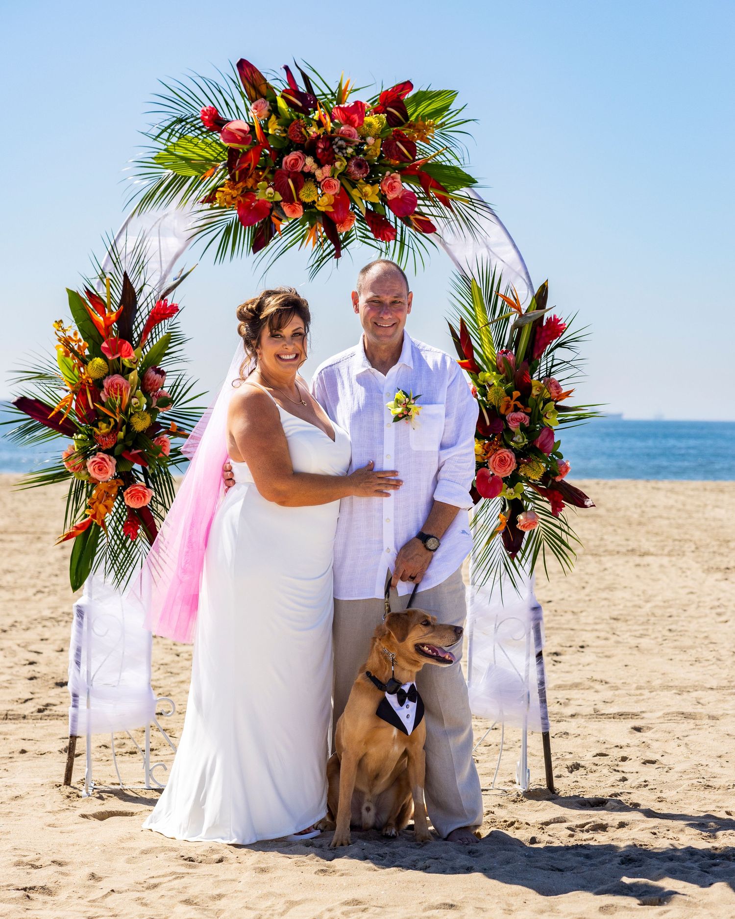 Orange County photographer captures Couple with a dog in a tuxedo posing under a tropical floral arch on a sunny beach.