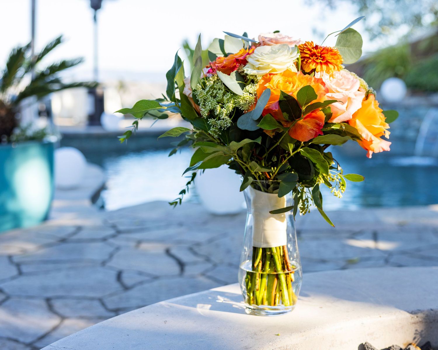 Orange County photographer captures Colorful orange and pink floral bouquet in a glass vase near a pool on a stone surface.