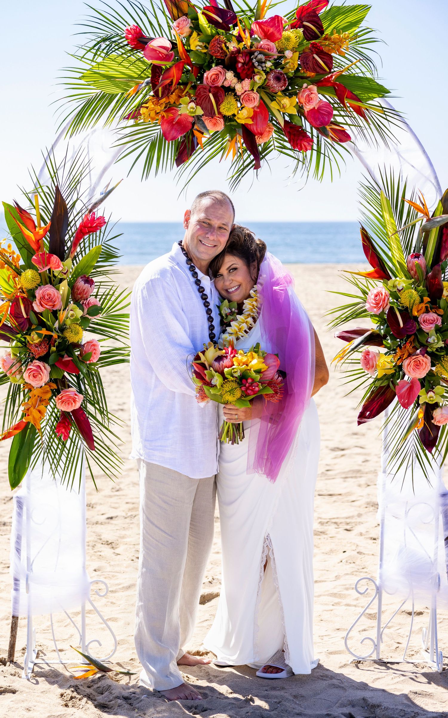 Orange County photographer captures Couple embracing under a vibrant tropical floral wedding arch on a sunny beach setting.