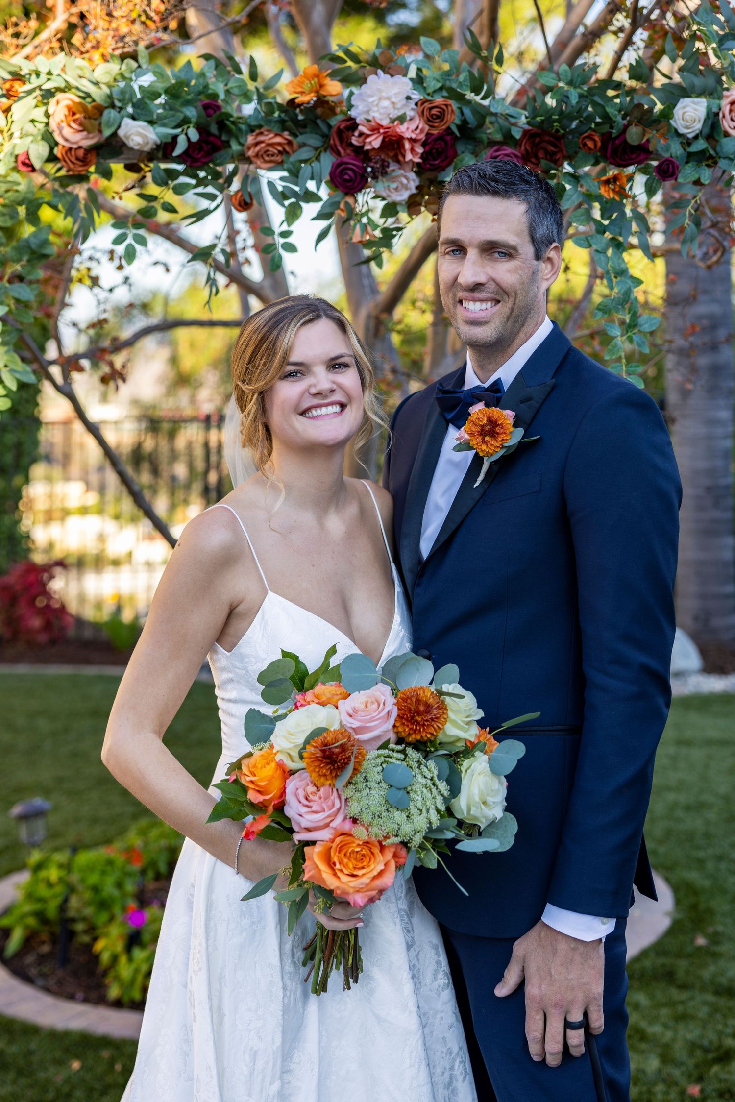 Orange County photographer captures Bride and groom smiling under a floral arch, bride holding an orange and pink bouquet outdoors.