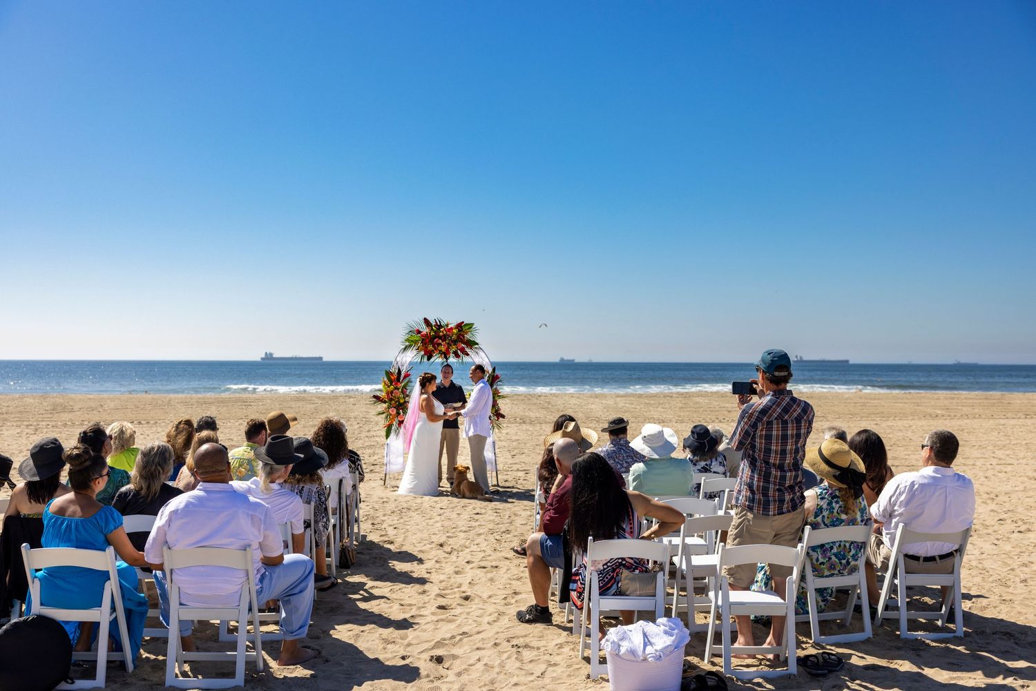 Orange County photographer captures Small beach wedding ceremony with guests seated on white chairs facing an arch by the ocean.