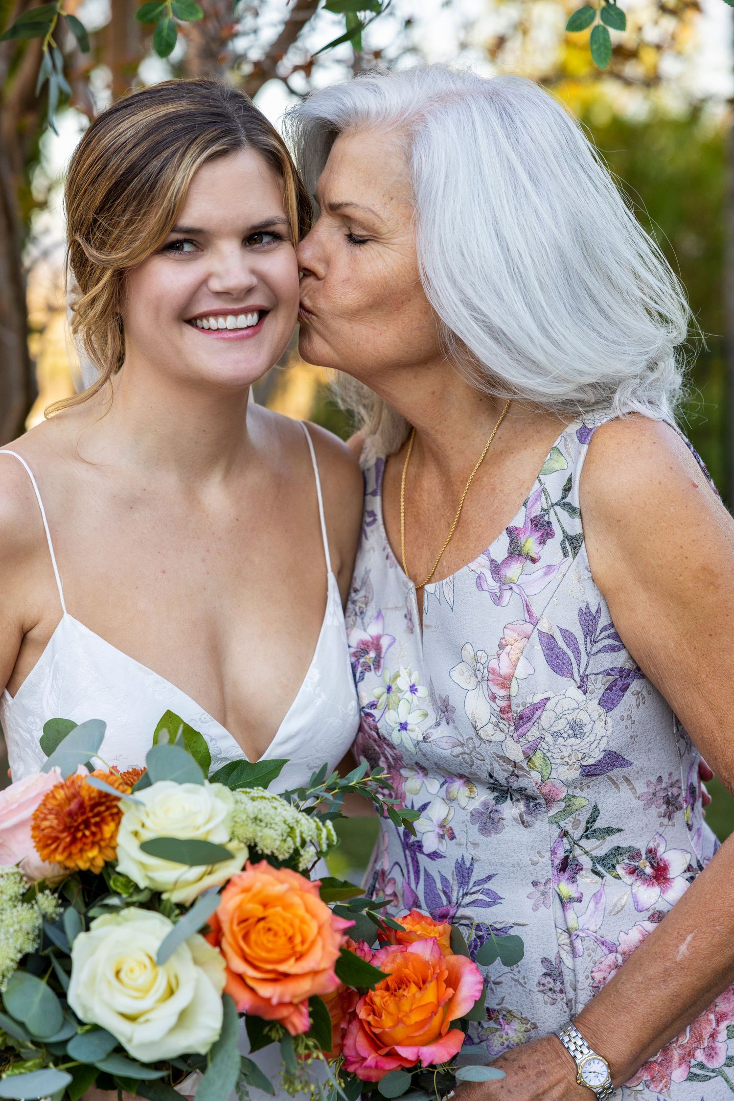Orange County photographer captures Smiling bride holding colorful bouquet receives a kiss on the cheek from an older woman outdoors.