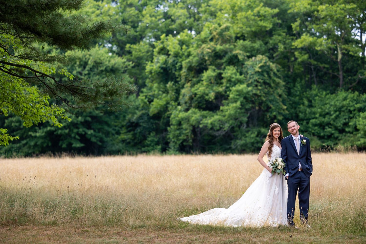 Wedding couple poses in rustic field with forest backdrop and flowing white dress.