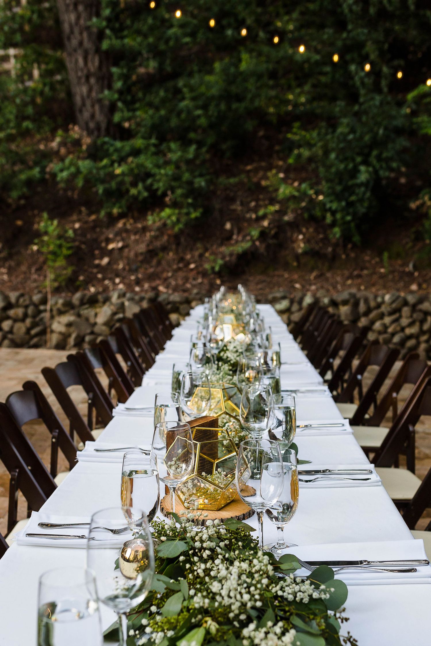 Long white banquet table with greenery runner and place settings in a forest setting with string lights.
