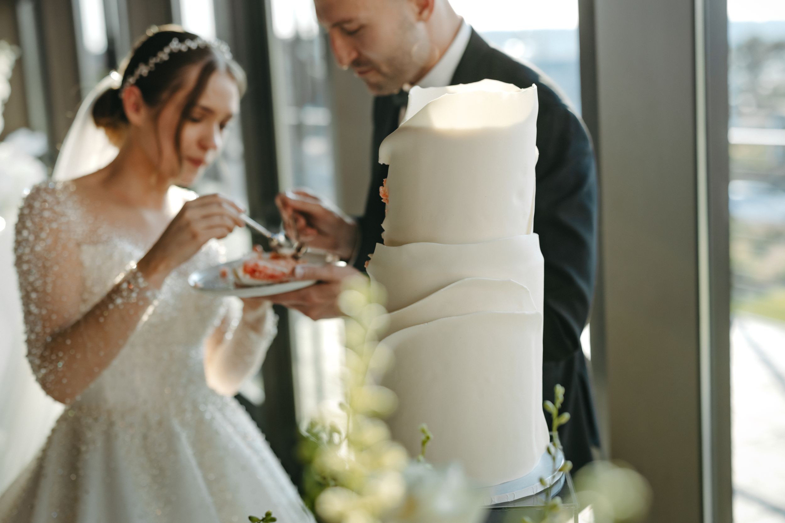 Wedding couple sharing cake during reception with white tiered cake and floral decor.