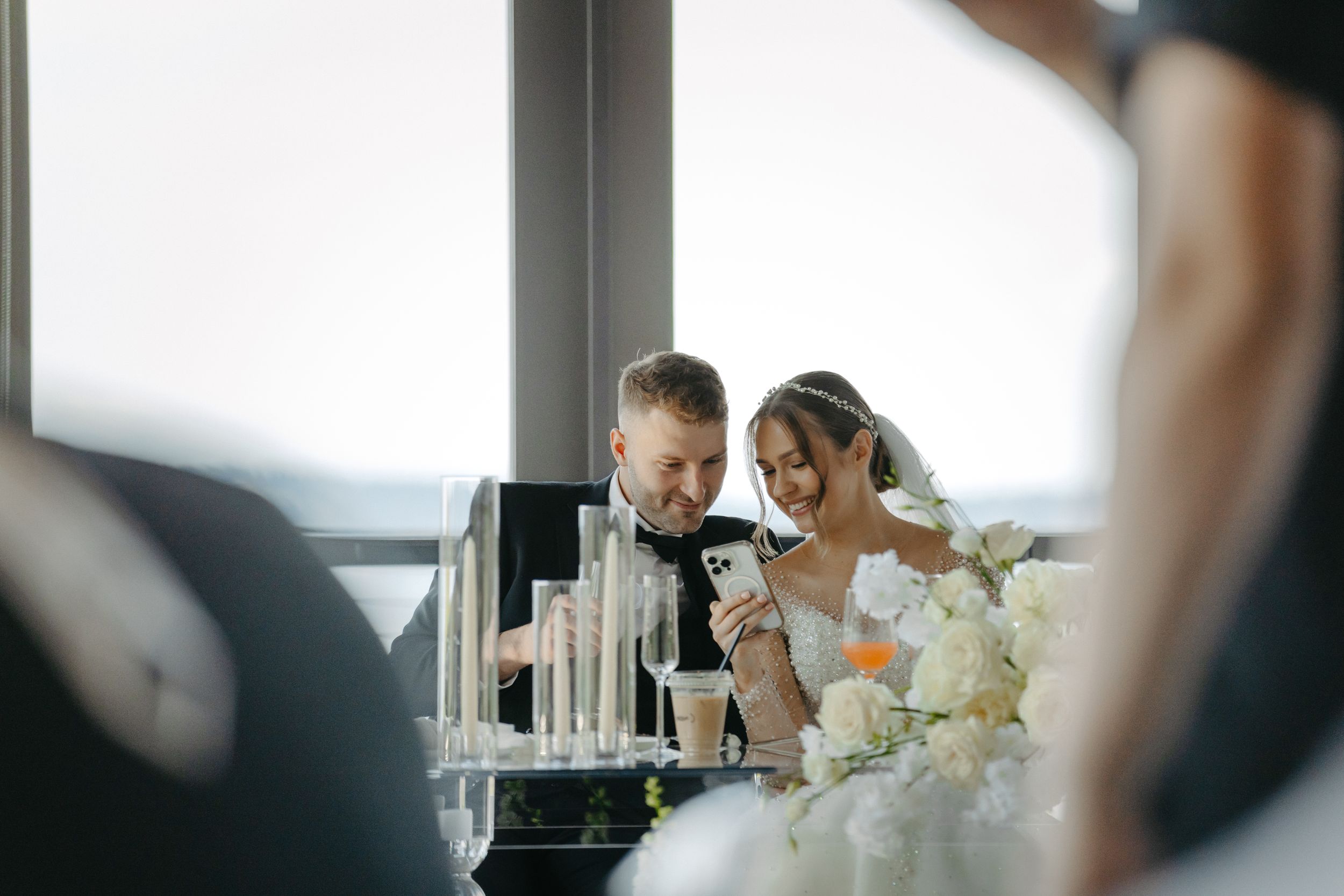 A wedding reception table set with white flowers and large windows overlooking water.