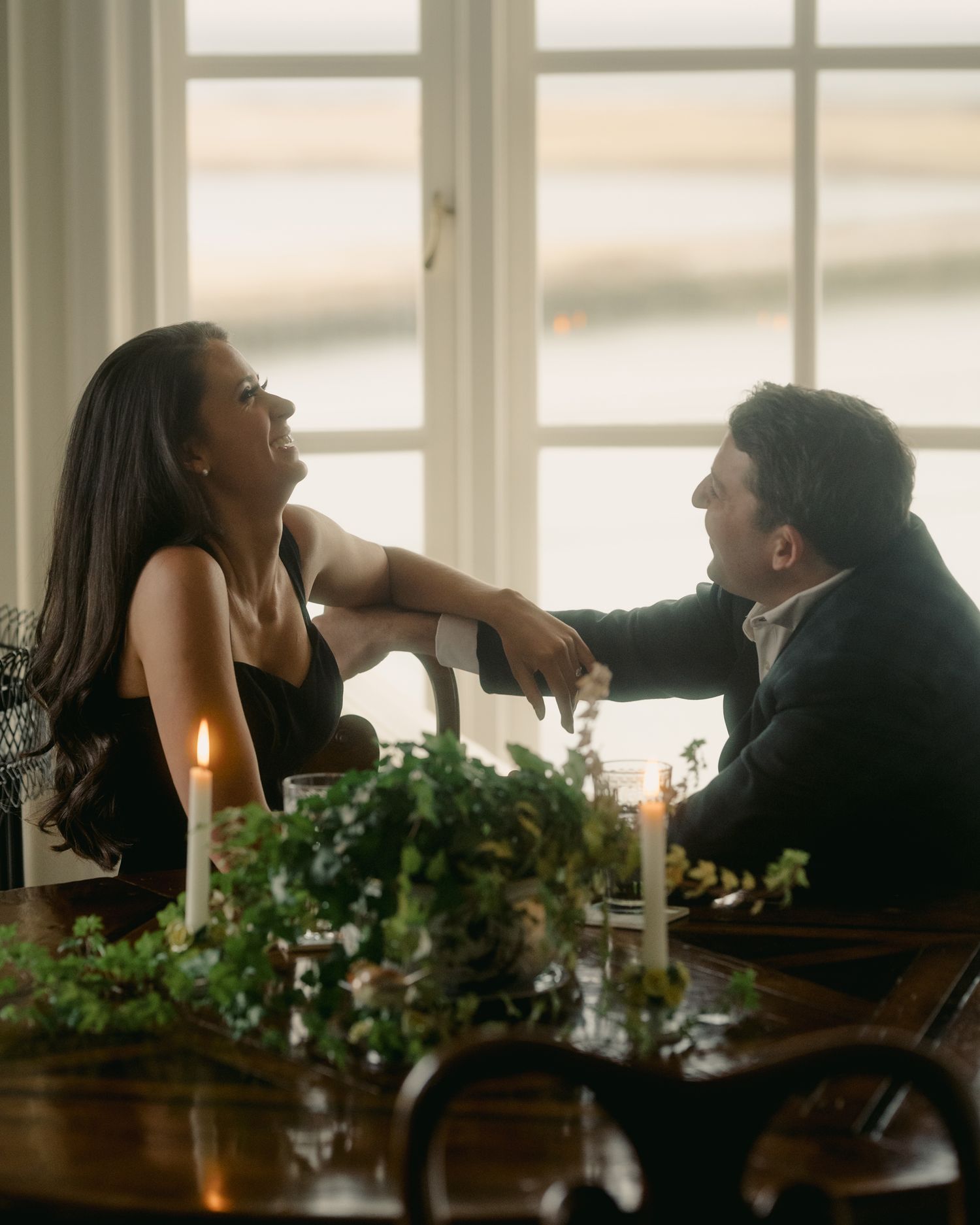 Romantic candlelit dinner scene with couple laughing at table decorated with greenery and white candles.
