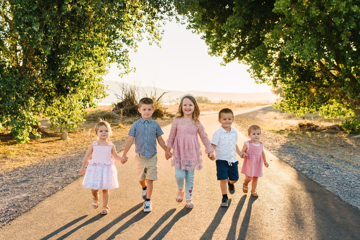 Children walking together on a dirt path with golden sunlight and green tree canopy overhead.